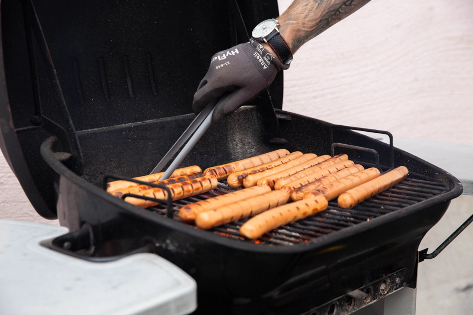 Det är en svart grill som står öppen, den har flera korvar i sig som är under tillagning. Det är en hand med handske på som håller i en tång som tar en av korvarna. 