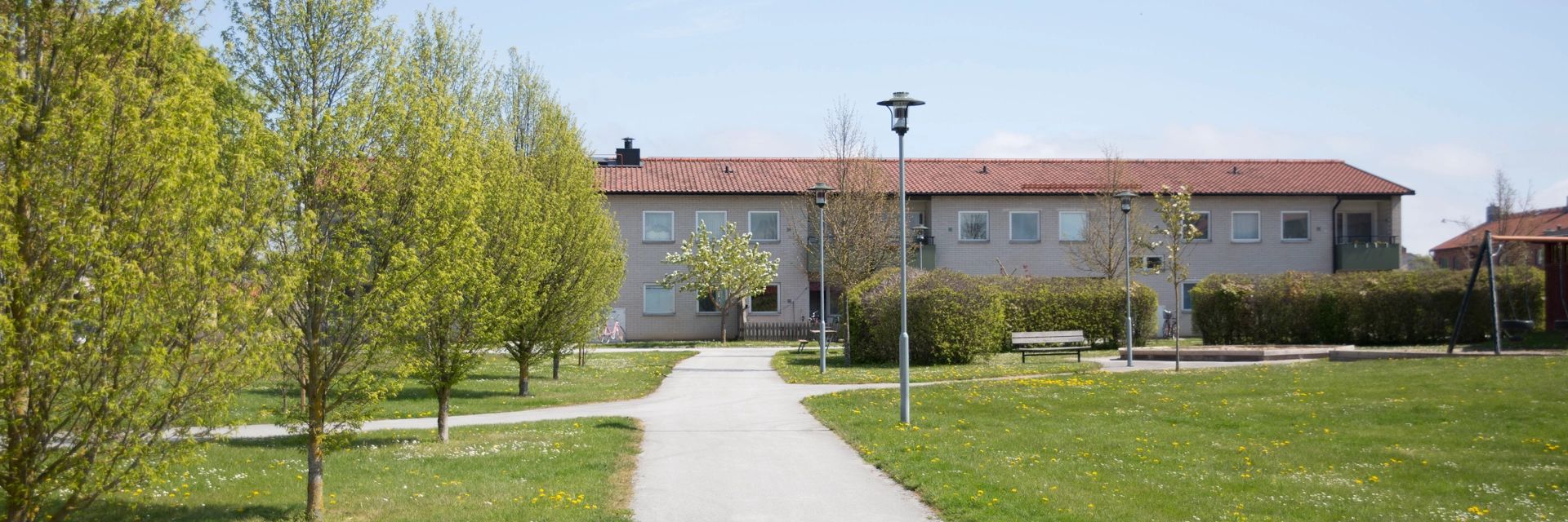 En gång väg med träd på ena sidan och en lekpark på andra sidan och i bakgrunden syns ett avlångt flerfamiljshus.