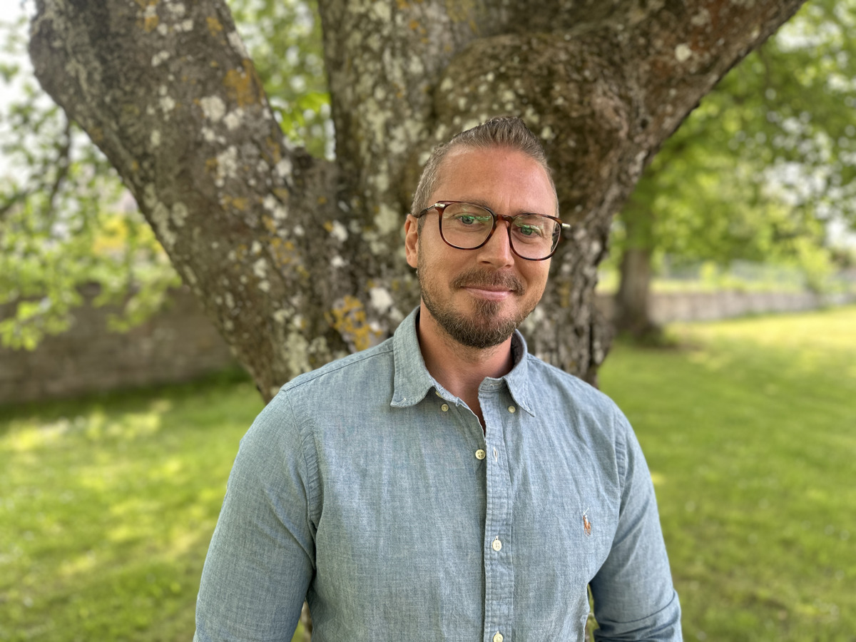 En man i blå skjorta och glasögon står framför ett träd och ler