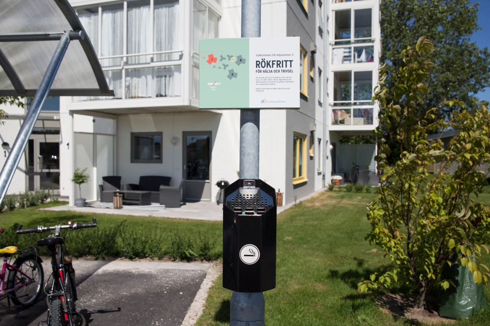 Skylt med texten "RÖKFRITT" på en lyktstolpe med askkopp. I bakgrunden syns ett vitt bostadshus med gula fönsterkarmar.