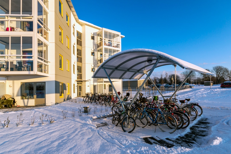 Solen skiner på ett flerbostadshus och det står cyklar under ett tak. Det är snö på marken.