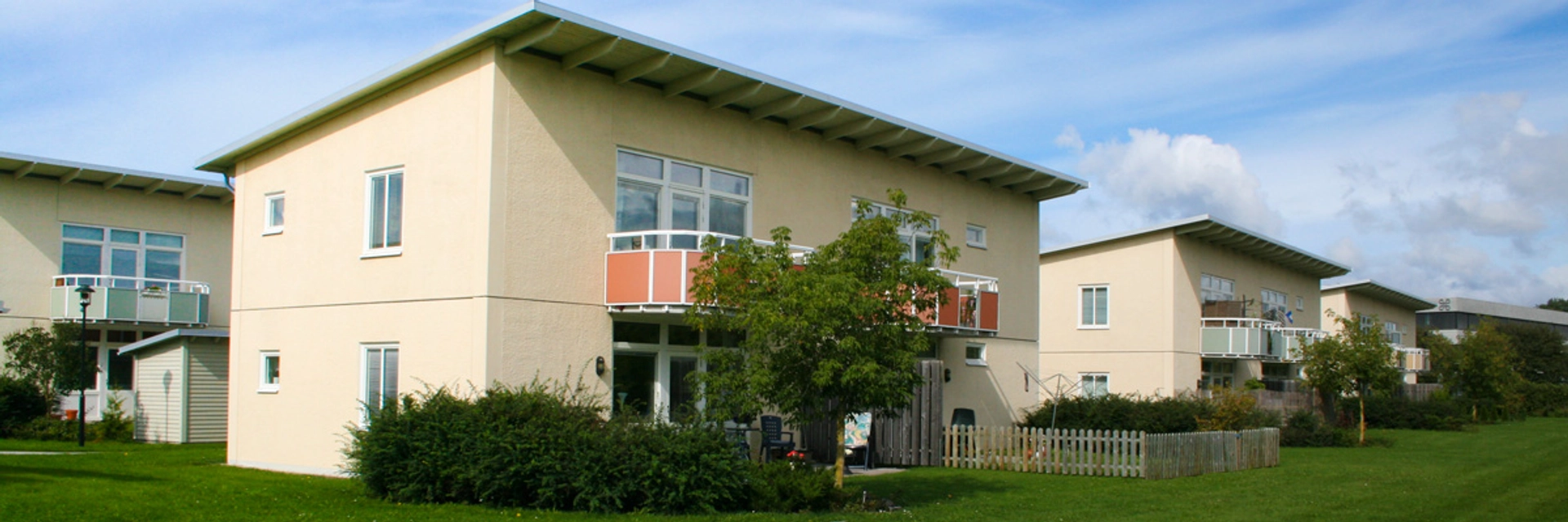 Tre tvåplans bostadshus med gul-beige fasad står på rad, ett till likadant hus syns bakom det första huset. Framför dem är grön gräsmatta och himlen är blå med flera moln.