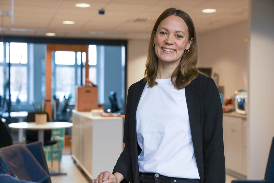 Kvinna med vit t-shirt och svart kofta står på ett kontor. Hon kollar in i kameran och ler. Det ser väldigt trevligt ut.
