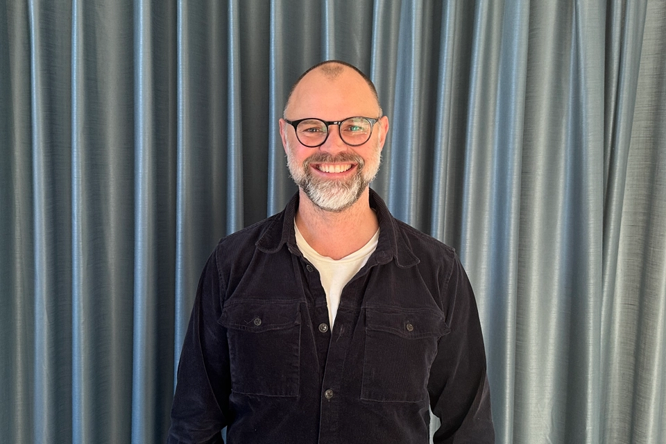 En man med svarta glasögon och skägg ser in i kameran och ler.