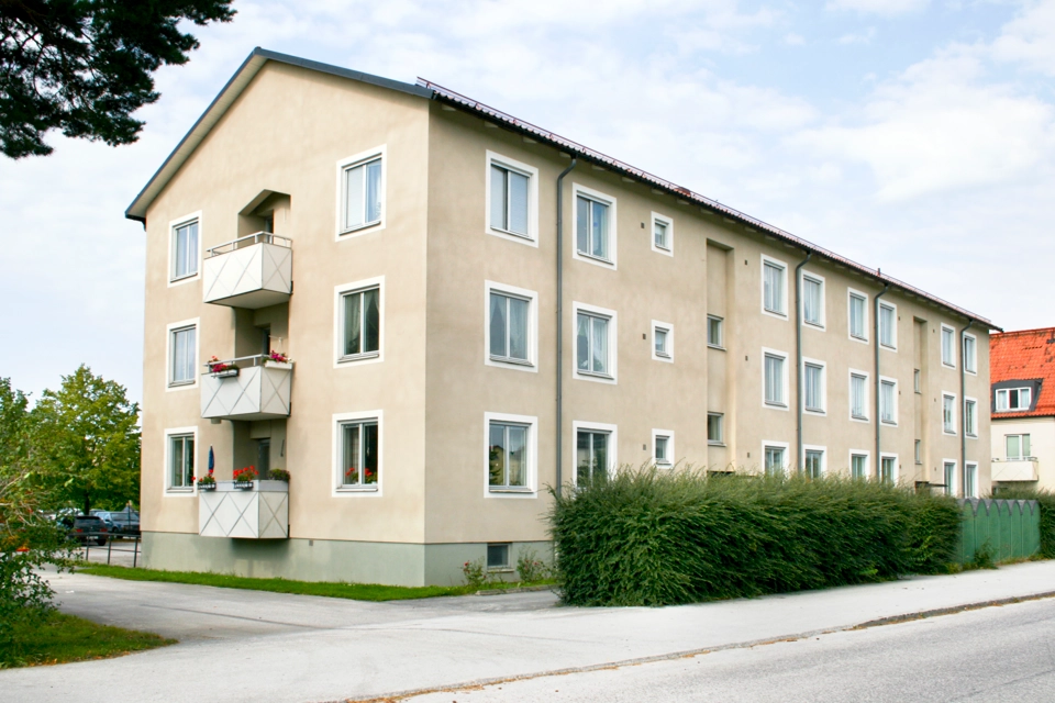 Treplans bostadshus med beige fasad, vita balkonger och vitt runt fönster. Framför huset går en häck och en trottoar. Det är flera moln på himlen.