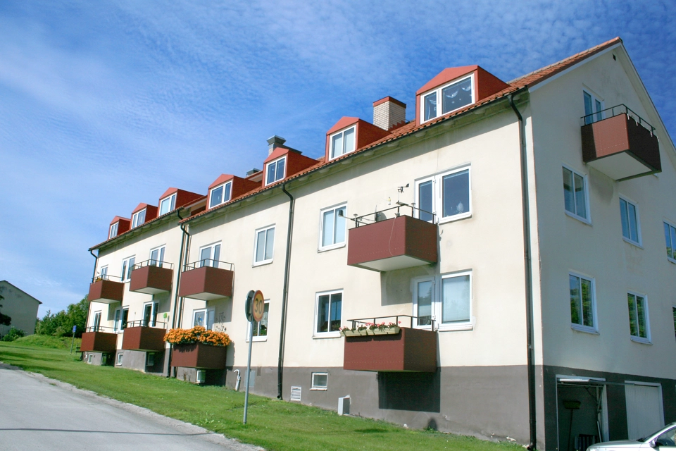 Vitt bostadshus med röda balkonger och rött tak. Framför huset är en gräsplätt. Framför gräset går en bilväg.