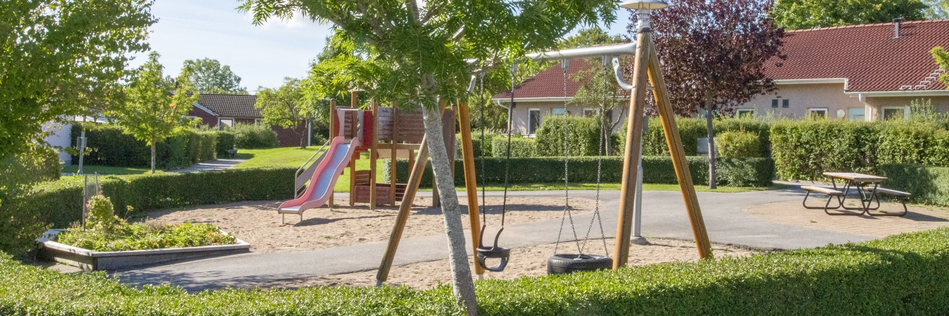 Lekplats i grönska, i bakgrunden skymtas en radhus med rött tegeltak