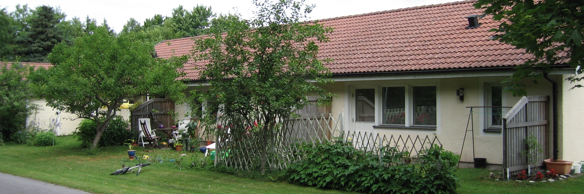 Enplanshus med ljusgul putsfasad och rött tegeltak. Längs huset finns två uteplatser separerade med ett plank. Framför dem är det gräsmatta och en smal, asfalterad gångväg.