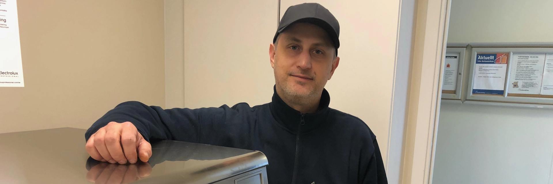 En man med svart keps och svart tröja med GotlandsHem loggan på står med armen på en tvättmaskin. 