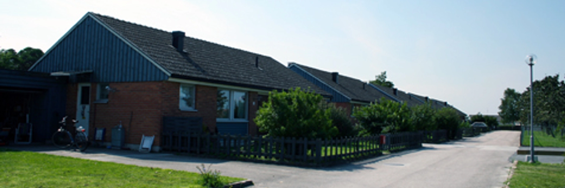 Rött tegelhus med blå träpanel på halva gavelsidan. Framför huset är en gräsplätt med staket omkring. Utanför går en asfalterad bilväg. Himlen är ljusblå.