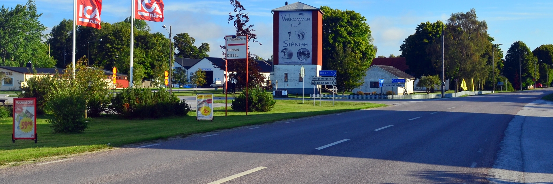 Flera skyltar och två röda flaggor med texten ICA står på en gräsmatta längs med en bilväg. Himlen är blå med ett fåtal moln.