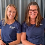 Två kvinnor i blå tröjor står  vid ett bord.