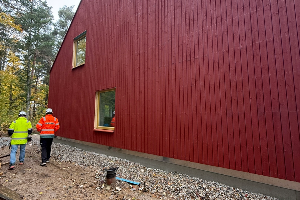 Två personer i varselkläder och bygghjälmar går bredvid ett nybyggt rött hus.