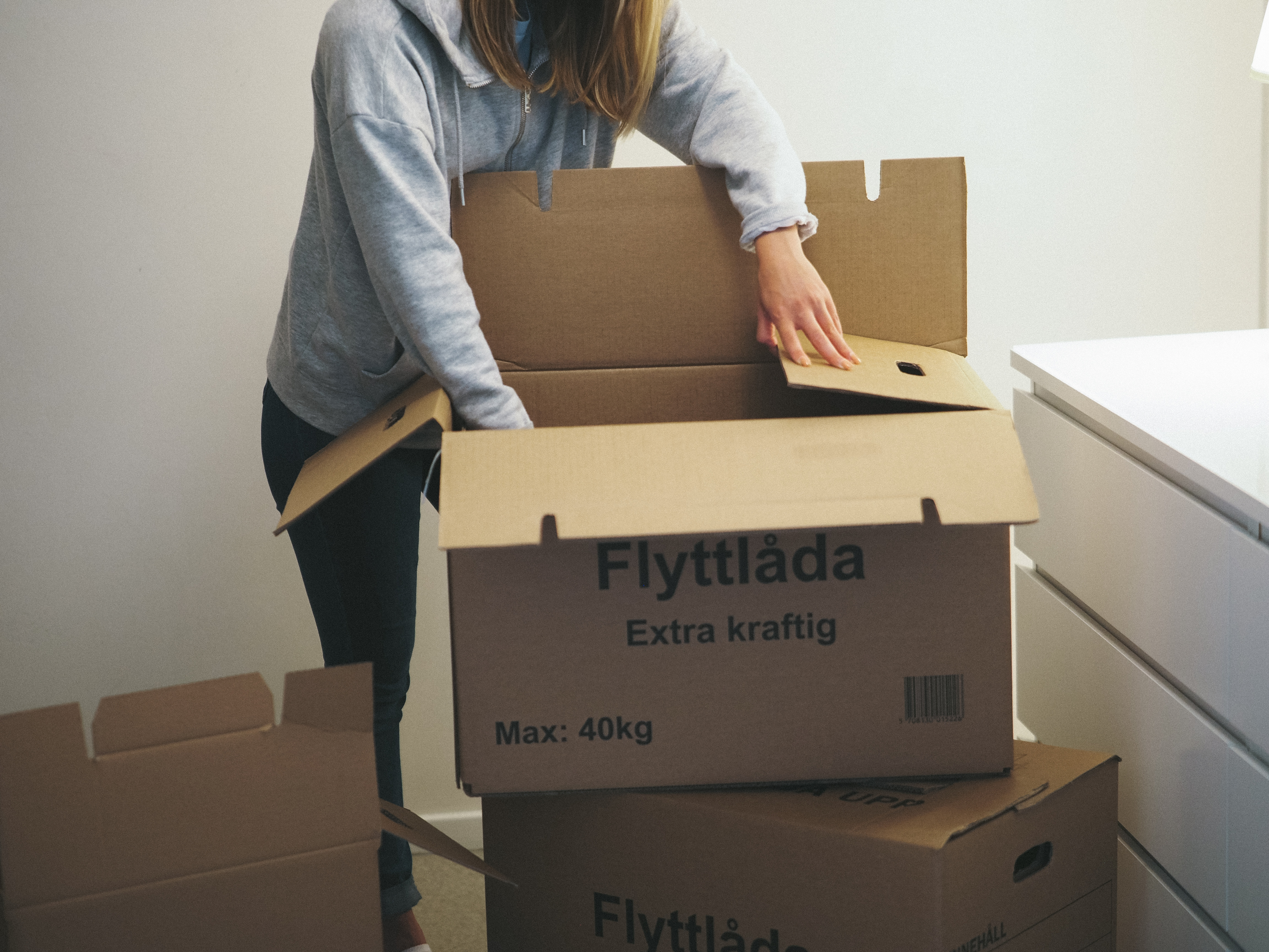 En kvinna med grå hoodie öppnar upp en flyttkartong inomhus.