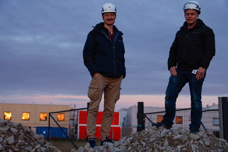 Två personer står på en grushög på en arbetsplats. Det är solnedgång.