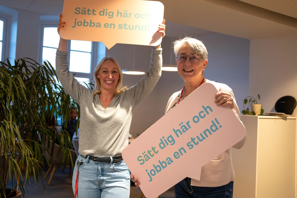 Två medarbetare ser glada ut och håller upp skyltar där det står "Sätt dig här och jobba en stund". Det ser inbjudande ut.