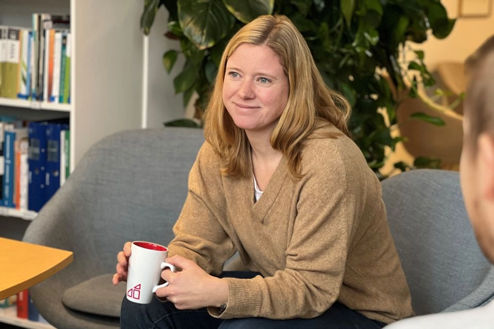 En kvinna med beige tröja sitter i en grå soffa, i handen håller hon en Gotlandshem mugg. Hon ser glad ut.